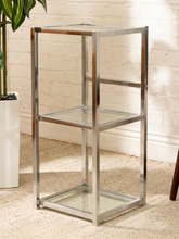 Load image into Gallery viewer, A three-tier metal and glass shelving unit with a polished chrome frame, standing on a beige woven rug against a white brick wall.