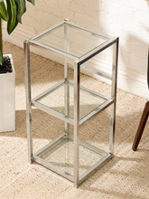 Load image into Gallery viewer, A three-tier side table with a polished chrome metal frame and clear glass shelves, shown in a styled room setting on a textured beige rug.