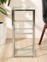 Load image into Gallery viewer, A three-tier side table with a chrome silver metal frame and clear glass shelves, shown in a styled room setting with a white brick wall, beige rug, and plant.