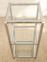 Load image into Gallery viewer, A modern two-tier side table featuring a polished silver metal frame and clear glass shelves, set against a neutral beige textured rug and white brick wall background.
