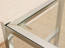 Load image into Gallery viewer, Close-up of a modern glass-top table corner with a brushed silver metal frame and Tempered Glass branding printed on the glass.