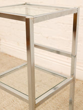 Load image into Gallery viewer, A modern two-tier side table featuring a polished silver metal frame with clear glass shelves and top, set against a white brick wall and beige carpet.