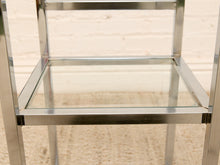 Load image into Gallery viewer, Close-up of a modern metal and glass side table or shelf with a brushed silver frame and clear glass panels, set against a light brick wall and beige carpet.