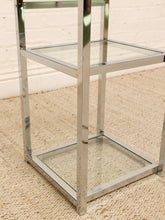 Load image into Gallery viewer, A modern two-tier side table with a polished chrome metal frame and clear glass shelves, shown on a beige textured rug against a white brick wall.