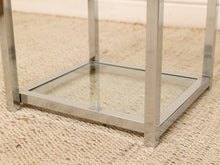 Load image into Gallery viewer, A close-up of a modern square side table with a brushed silver metal frame and a clear glass bottom shelf, set on a beige woven rug.