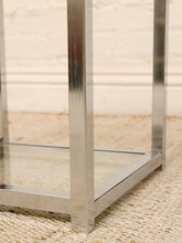 Load image into Gallery viewer, Close-up of a modern side table frame with polished silver metal legs and a glass shelf, set on a woven beige rug against a light brick wall.
