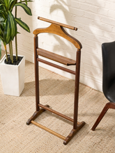 Load image into Gallery viewer, A wooden clothes rack with a curved top bar and two horizontal support bars, shown standing on a textured beige rug against a white brick wall, next to a potted plant and a chair.