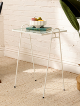 Load image into Gallery viewer, A small white metal side table with a clear glass top, styled with a bowl of fruit and a book, set against a white brick wall on a beige rug.