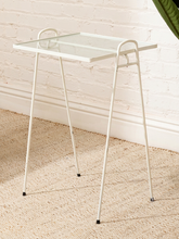Load image into Gallery viewer, A white metal side table with a clear glass tray top and curved legs, shown in a room setting on a beige woven rug against a white brick wall.