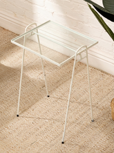 Load image into Gallery viewer, A modern white metal side table with a clear glass top, set on a textured beige jute rug against a white brick wall.