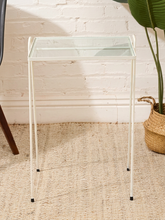 Load image into Gallery viewer, A modern white metal side table with a clear glass top, standing on a textured beige rug against a white brick wall, with a potted plant and part of a chair visible in the background.