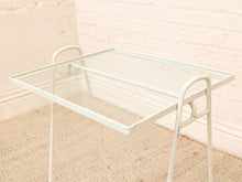 Load image into Gallery viewer, A white metal side table with a rectangular clear glass top and integrated side handles, set against a white brick wall and beige carpet.