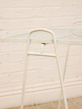 Load image into Gallery viewer, A close-up of a white metal table frame with a clear glass tabletop, set against a white brick wall background.