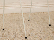 Load image into Gallery viewer, Close-up of white metal hair stand legs with black rubber feet resting on a beige woven rug against a white wall.