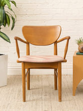 Load image into Gallery viewer, A mid-century modern wooden armchair with a curved backrest and a light pink upholstered seat, set against a white brick wall with a small wooden side table and potted plants.