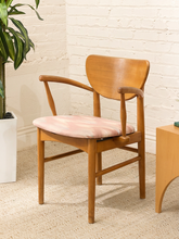Load image into Gallery viewer, A mid-century modern wooden armchair with a curved backrest, armrests, and a pink and cream patterned upholstered seat cushion, shown in a room setting with a white brick wall, beige carpet, and a potted plant.