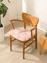 Load image into Gallery viewer, A mid-century modern wooden armchair with a curved backrest and armrests, featuring a light pink and white abstract patterned seat cushion, set in a bright room with a white brick wall and beige carpet.