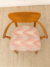 Load image into Gallery viewer, A wooden dining chair with a curved backrest and armrests, featuring a pink, beige, and white patterned seat cushion, shown on a woven jute rug.