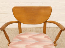 Load image into Gallery viewer, A mid-century modern wooden armchair with a curved backrest and upholstered seat in a pink, beige, and white chevron pattern, set against a white brick wall and textured rug.