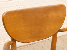 Load image into Gallery viewer, A close-up of a wooden chair backrest with a warm medium-brown finish, set against a white painted brick wall and light beige carpet.