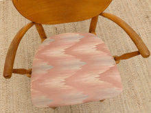 Load image into Gallery viewer, A mid-century modern wooden armchair with a pink, beige, and light blue ikat patterned upholstered seat cushion, shown from a top-down angle on a woven beige rug.