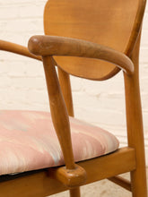 Load image into Gallery viewer, A close-up of a mid-century modern wooden armchair with a curved backrest, turned wooden armrests, and a pink and white striped upholstered seat cushion, set against a white brick wall.
