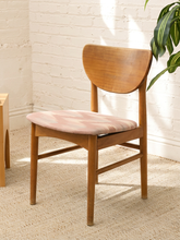 Load image into Gallery viewer, A mid-century modern wooden dining chair with a curved backrest and a blush pink and white marbled upholstered seat cushion, set against a white brick wall with a plant leaf in the corner.
