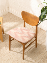 Load image into Gallery viewer, A mid-century modern wooden dining chair with a light pink and beige ikat-patterned cushion, set on a textured beige rug in a bright room.