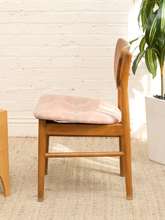 Load image into Gallery viewer, A wooden dining chair with a light pink cushioned seat and a matching pink backrest, set against a white brick wall and a beige rug next to a potted plant.