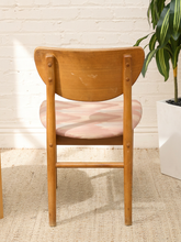 Load image into Gallery viewer, A mid-century modern wooden dining chair with a curved backrest, natural oak frame, and a pink and white patterned seat cushion, shown in a room setting with a white brick wall and a potted plant.