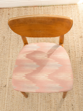 Load image into Gallery viewer, A mid-century modern wooden chair viewed from above, featuring a warm medium-brown wood frame and a pink, beige, and white zigzag patterned seat cushion, set on a woven beige rug.