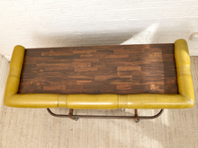 Load image into Gallery viewer, A rectangular bench with a dark brown wood plank top and mustard yellow leather padded arms and sides, supported by a dark metal frame, photographed against a white brick wall and light rug.