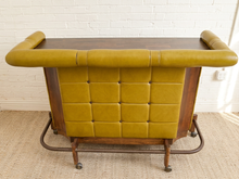 Load image into Gallery viewer, A vintage bar cart with a mustard yellow tufted leather front and backrest, a dark wood top, and a metal rolling frame, set against a white brick wall and beige carpet.