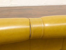 Load image into Gallery viewer, Close-up of a mustard yellow leather sofa armrest showing the stitched seam and texture, with a dark wood surface and white brick wall in the background.