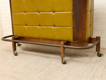 Load image into Gallery viewer, A mid-century modern rolling storage cabinet with mustard yellow tufted leather upholstery, a dark wood frame, and a curved metal base on casters, shown on a beige carpet against a white brick wall.