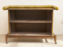 Load image into Gallery viewer, A mid-century modern rolling storage cart with a dark brown wood frame, two open shelves, and mustard yellow tufted leather upholstery on the top surface and sides, set against a white brick wall.