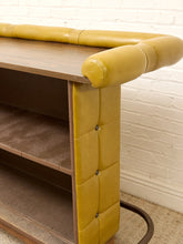 Load image into Gallery viewer, A close-up of a mid-century modern console table featuring a warm mustard yellow tufted leather upholstery and a medium brown wood grain top and shelves, set against a white brick wall.