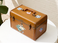 Load image into Gallery viewer, A vintage brown leather suitcase with floral and bird illustrations, displayed on a white round table against a white brick wall.