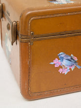 Load image into Gallery viewer, A close-up of a vintage brown leather suitcase corner decorated with a blue bird and pink flower sticker, resting on a light fabric surface.
