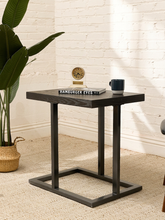 Load image into Gallery viewer, A dark wood and metal side table with a square top and open rectangular base, styled in a bright room with a white brick wall, beige rug, potted plant, and small tabletop decor.