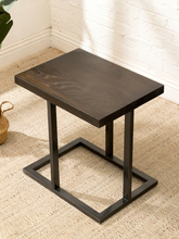 Load image into Gallery viewer, A square dark wood side table with a black metal frame base, set on a beige woven rug against a white brick wall.