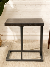 Load image into Gallery viewer, A dark wood and metal side table with a rectangular top and a square open-frame base, shown in a styled interior setting with a white brick wall and woven rug.