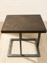 Load image into Gallery viewer, A square dark brown wood end table with a dark gray metal frame base, set on a woven jute rug against a white brick wall.