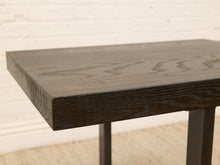 Load image into Gallery viewer, Close-up of a dark stained wooden table corner showing the wood grain and edge finish, with dark metal legs visible against a light wall and woven floor.