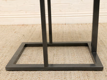 Load image into Gallery viewer, A close-up of a dark charcoal gray metal table base with a square frame and vertical support legs, resting on a beige woven rug against a white brick wall.