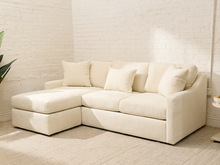 Load image into Gallery viewer, An off-white upholstered sectional sofa with a chaise lounge on the left, styled with matching throw pillows in a bright, modern room with a white brick wall and light beige rug.