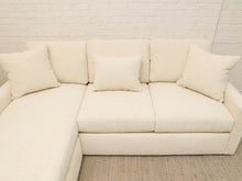 Load image into Gallery viewer, An off-white textured fabric sectional sofa with matching throw pillows, set against a white brick wall and a woven rug.