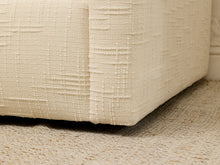 Load image into Gallery viewer, Close-up of a cream-colored upholstered furniture corner with a textured basketweave fabric, resting on a woven beige rug.
