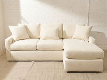 Load image into Gallery viewer, An off-white textured fabric sectional sofa with a right-facing chaise lounge and four matching throw pillows, set against a white brick wall and a light beige woven rug.