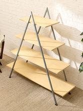 Load image into Gallery viewer, A modern A-frame shelving unit with four light natural wood shelves and gray metal legs, set against a white brick wall on a beige carpet.